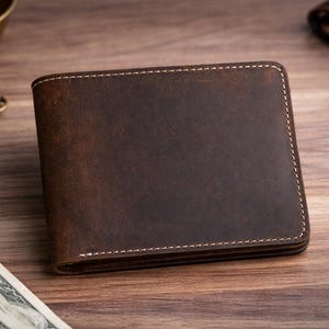 May include: A brown leather wallet with white stitching. The wallet is rectangular and appears to be new. Keys, a small bowl, a watch, and cash are also on the wooden surface.