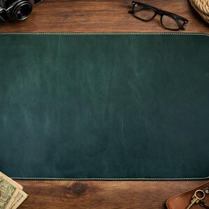 May include: A dark green leather desk mat with white stitching, surrounded by a vintage camera, glasses, a pen, a cup of coffee, a wallet, and other accessories on a wooden surface.