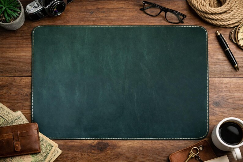 May include: A dark green leather desk mat with white stitching, surrounded by a vintage camera, glasses, a pen, a cup of coffee, a wallet, and other accessories on a wooden surface.