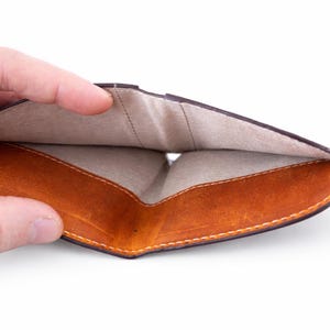 K&ouml;nnte beinhalten: Ge&ouml;ffnetes braunes Lederportemonnaie mit hellem, beigem Innenraum. Das Portemonnaie hat ein geschwungenes Design und wird offen gehalten, wodurch die Innenf&auml;cher und die Nahtdetails zur Geltung kommen. Das Leder hat einen Used-Look.