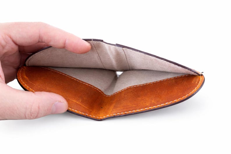 K&ouml;nnte beinhalten: Ge&ouml;ffnetes braunes Lederportemonnaie mit hellem, beigem Innenraum. Das Portemonnaie hat ein geschwungenes Design und wird offen gehalten, wodurch die Innenf&auml;cher und die Nahtdetails zur Geltung kommen. Das Leder hat einen Used-Look.