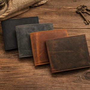 May include: Four leather wallets in black, grey, brown, and dark brown are arranged on a wooden surface. The wallets are rectangular and appear to be bi-fold. The image also includes keys, stones, and a rope.