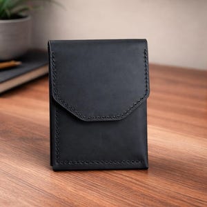 May include: A black leather wallet with a flap closure, featuring detailed stitching along the edges. The wallet is rectangular and stands upright on a wooden surface. The background includes a notebook, pen, and a potted plant.