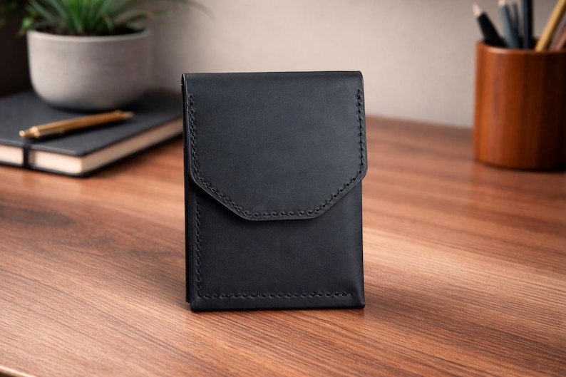May include: A black leather wallet with a flap closure, featuring detailed stitching along the edges. The wallet is rectangular and stands upright on a wooden surface. The background includes a notebook, pen, and a potted plant.