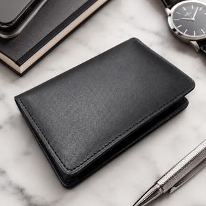 May include: A rectangular black leather wallet with visible stitching along the edges. The wallet is positioned centrally, accompanied by a black notebook, a silver-toned wristwatch with a black leather strap, and a metallic silver pen.