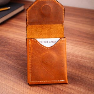May include: Open leather card holder in a warm brown hue, with visible stitching and a snap closure. The interior reveals a card with the words "CREDIT CARD." The wallet is set against a wooden surface, showcasing its texture and craftsmanship.