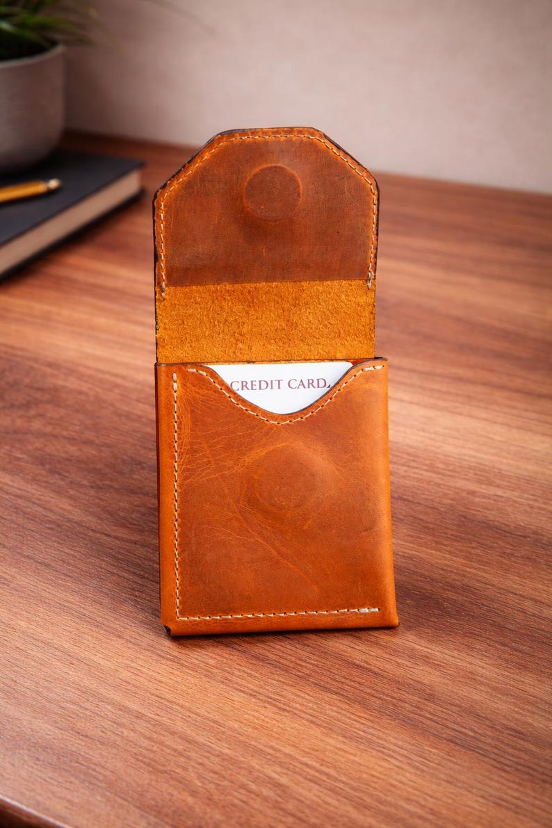 May include: Open leather card holder in a warm brown hue, with visible stitching and a snap closure. The interior reveals a card with the words "CREDIT CARD." The wallet is set against a wooden surface, showcasing its texture and craftsmanship.