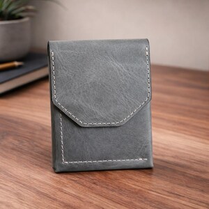 May include: A gray leather wallet with a flap closure and white stitching. The wallet is rectangular with a geometric flap design. It is sitting on a wooden surface.