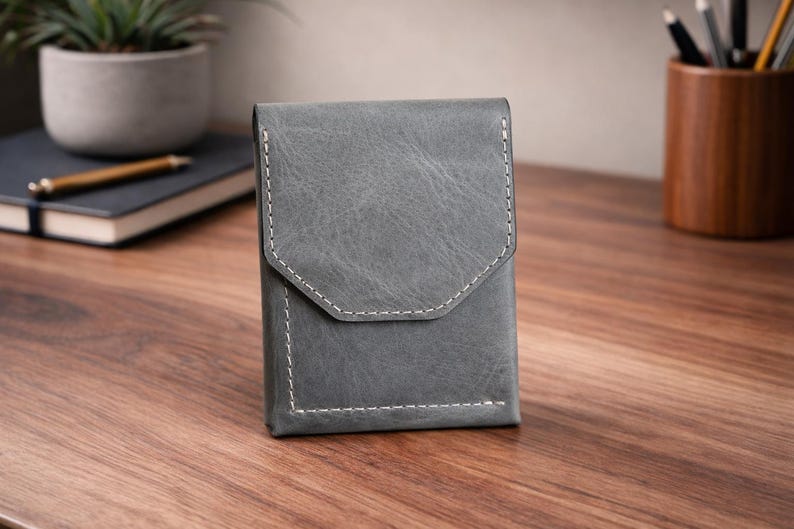 May include: A gray leather wallet with a flap closure and white stitching. The wallet is rectangular with a geometric flap design. It is sitting on a wooden surface.