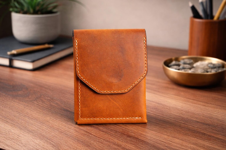 May include: A rectangular, brown leather wallet with a flap closure. The wallet is made of textured leather and has visible stitching. It is sitting on a wooden surface. The background includes a small potted plant, a notebook, and a pen.