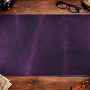 May include: A rectangular, deep purple leather desk mat with white stitching. The mat is surrounded by a wooden desk, a cup of coffee, a camera, glasses, and other accessories.