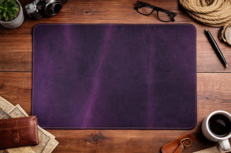 May include: A rectangular, deep purple leather desk mat with white stitching. The mat is surrounded by a wooden desk, a cup of coffee, a camera, glasses, and other accessories.