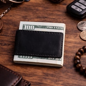 Op de afbeelding: Een zwarte leren geldclip met een stapel Amerikaanse honderd dollar biljetten. De geldclip is rechthoekig met een gestikte rand. Andere items op het houten oppervlak zijn een portemonnee, zonnebril en munten.
