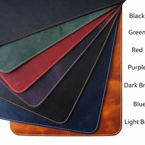 May include: A fan of leather desk pads in various colors, including black, green, red, purple, dark brown, blue, and light brown. Each pad has a contrasting stitch around the edge. The colors are labeled on the right side of the image.