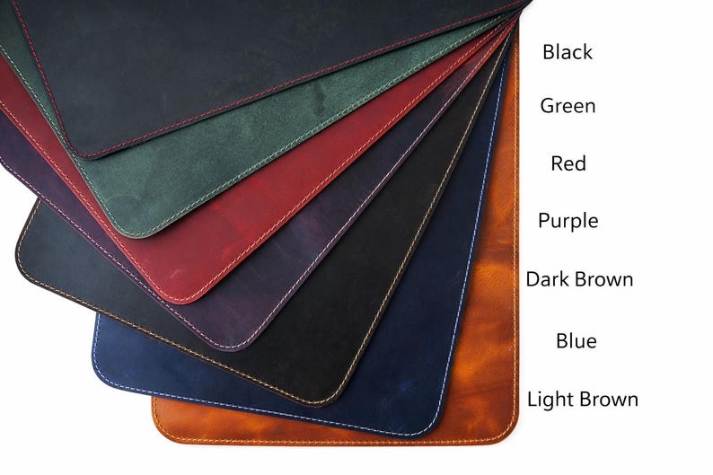 May include: A fan of leather desk pads in various colors, including black, green, red, purple, dark brown, blue, and light brown. Each pad has a contrasting stitch around the edge. The colors are labeled on the right side of the image.
