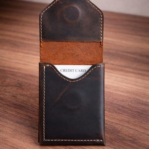 May include: A dark brown leather card holder with white stitching and a curved top flap. The interior is a lighter brown, and a white card with "CREDIT CARD" printed on it is visible.