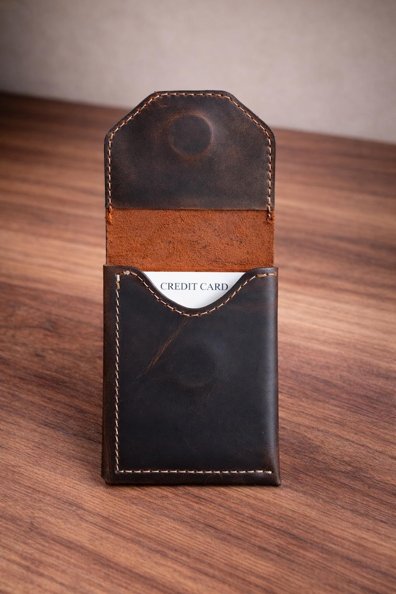 May include: A dark brown leather card holder with white stitching and a curved top flap. The interior is a lighter brown, and a white card with "CREDIT CARD" printed on it is visible.