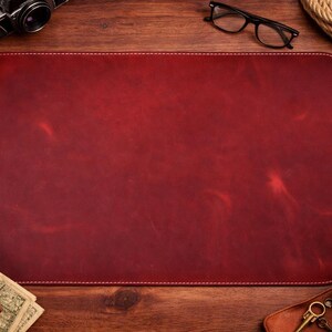 May include: A rectangular, burgundy leather desk mat with white stitching. The mat is surrounded by a wooden desk, a cup of coffee, a wallet, a camera, and other accessories.