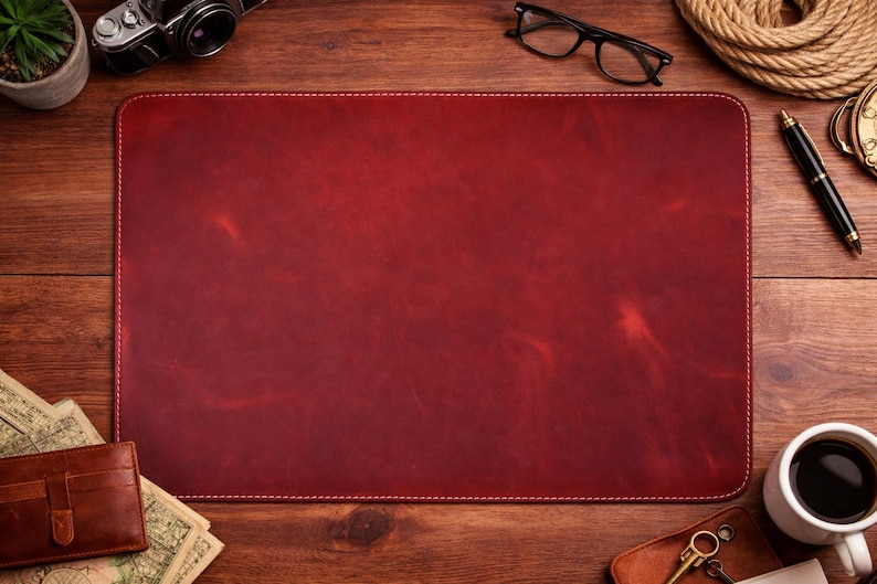 May include: A rectangular, burgundy leather desk mat with white stitching. The mat is surrounded by a wooden desk, a cup of coffee, a wallet, a camera, and other accessories.