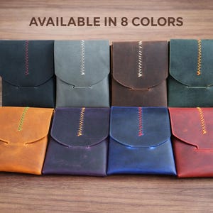 May include: A collection of eight leather wallets in various colors, including black, gray, brown, and red. Each wallet features a flap closure and a decorative stitched detail. The text "AVAILABLE IN 8 COLORS" is at the top.