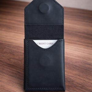 May include: Black leather card holder with a snap closure. The interior reveals a white card with the words "CREDIT CARD". The wallet is open, showcasing its storage capacity. The stitching is visible along the edges.