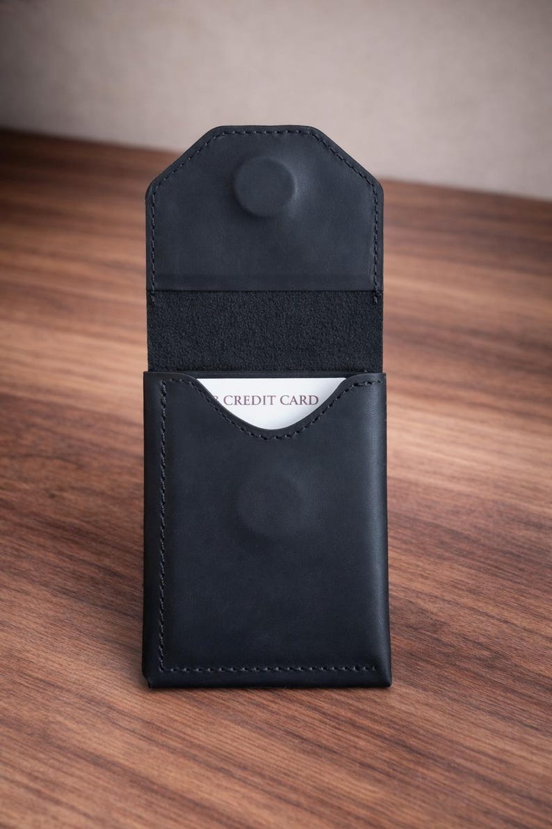 May include: Black leather card holder with a snap closure. The interior reveals a white card with the words "CREDIT CARD". The wallet is open, showcasing its storage capacity. The stitching is visible along the edges.