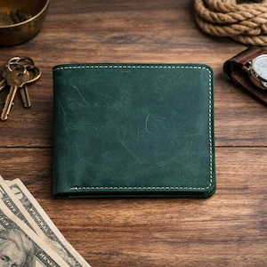 May include: A dark teal leather wallet with white stitching. The wallet is rectangular and sits on a wooden surface, with keys, cash, and a watch visible in the background. The wallet has a distressed finish.