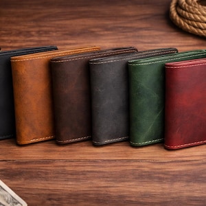 May include: A collection of six leather wallets in various colors, including black, brown, green, and red. Each wallet is rectangular with visible stitching and a textured surface. The wallets are arranged in a row on a wooden surface.