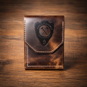 May include: A brown leather wallet with a flap closure. The wallet features a stitched design and a black emblem with the text "MEN'S ALLIANCE" and "BIRD DOG". The wallet is made of distressed leather.