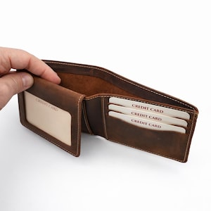 May include: Open brown leather wallet with multiple card slots. The wallet has a clear ID window and slots holding three credit cards. The interior is a lighter brown, and the stitching is visible.