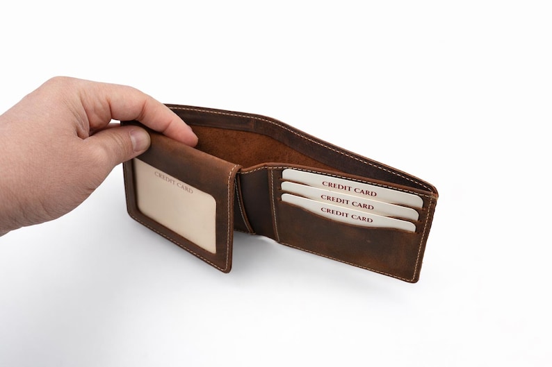 May include: Open brown leather wallet with multiple card slots. The wallet has a clear ID window and slots holding three credit cards. The interior is a lighter brown, and the stitching is visible.