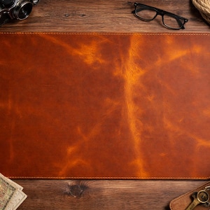 Pode incluir: Um tapete de mesa retangular de couro marrom com uma superfície texturizada. O tapete é cercado por uma mesa de madeira, uma câmera, óculos, uma caneta, uma carteira, uma xícara de café e outros acessórios.