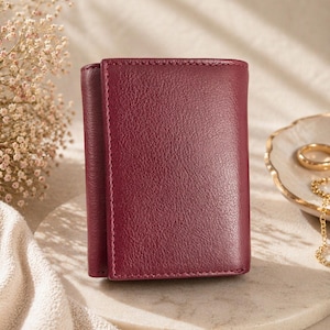 May include: A burgundy leather wallet with a tri-fold design. The wallet is made of textured leather and features visible stitching around the edges. The wallet is displayed on a round, light-colored surface.
