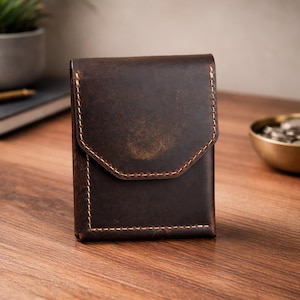 May include: A dark brown leather wallet with a flap closure and contrasting stitching. The wallet is rectangular and appears to be made of distressed leather. The background includes a desk, a notebook, and a small plant.