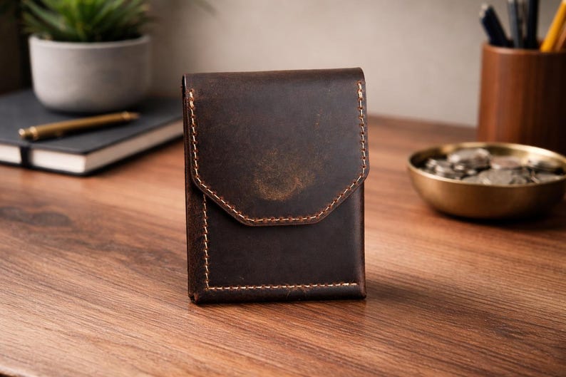 May include: A dark brown leather wallet with a flap closure and contrasting stitching. The wallet is rectangular and appears to be made of distressed leather. The background includes a desk, a notebook, and a small plant.