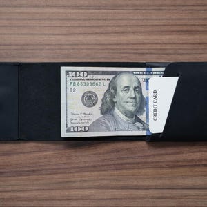 May include: A black leather wallet, open to display a one hundred dollar bill and a credit card. The wallet is on a wooden surface. The credit card is white with the words "CREDIT CARD" printed on it.
