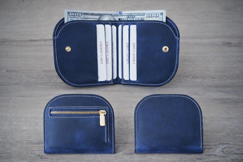 K&ouml;nnte beinhalten: Ein marineblaues Lederportemonnaie mit wei&szlig;en N&auml;hten, ge&ouml;ffnet, um Kreditkarten und einen Stapel 100-Dollar-Scheine zu zeigen. Zwei weitere Geldb&ouml;rsen sind abgebildet, eine mit Rei&szlig;verschluss und goldfarbener Hardware und eine geschlossene.