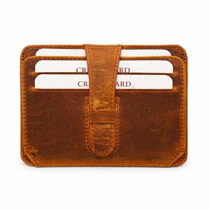 May include: A brown leather card wallet with a flap closure. The wallet has a worn, textured appearance and holds several white cards. The cards are partially visible, with the word "CARD" printed on them. The wallet is rectangular and has rounded corners.