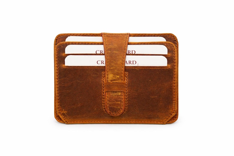 May include: A brown leather card wallet with a flap closure. The wallet has a worn, textured appearance and holds several white cards. The cards are partially visible, with the word "CARD" printed on them. The wallet is rectangular and has rounded corners.