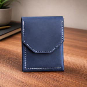 May include: A navy blue leather wallet with white stitching. The wallet has a flap closure with a unique geometric design. The product is displayed on a wooden surface.