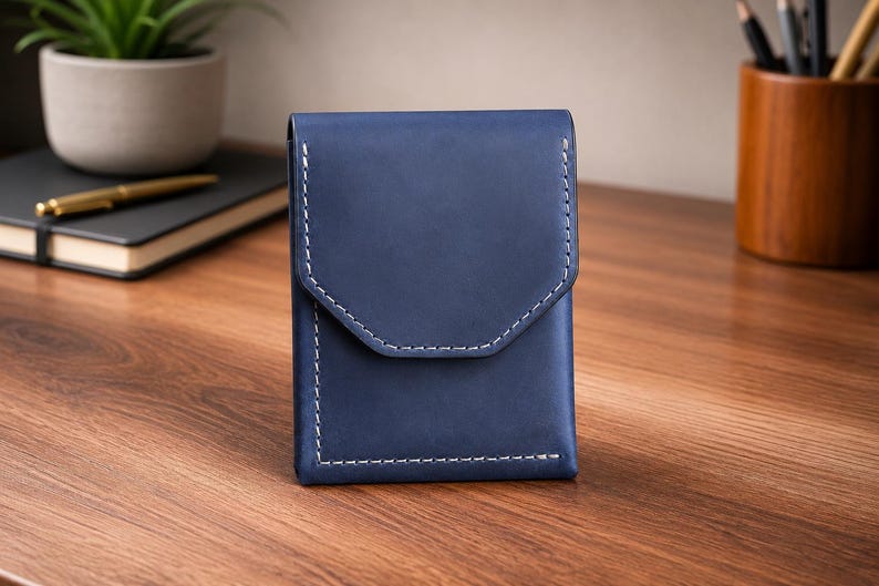 May include: A navy blue leather wallet with white stitching. The wallet has a flap closure with a unique geometric design. The product is displayed on a wooden surface.