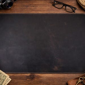 May include: A black leather desk mat with white stitching, surrounded by office supplies on a wooden surface. Items include a camera, glasses, a pen, a cup of coffee, and a wallet with cash.