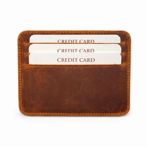 May include: A brown leather card holder with three white credit cards visible. The cards are labeled "CREDIT CARD" in a bold, red font. The card holder has a rectangular shape with rounded corners and a stitched border.