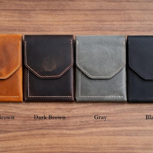 May include: Four leather wallets in light brown, dark brown, gray, and black, arranged on a wooden surface. Each wallet has a flap closure and visible stitching. The colors are labeled below each wallet.
