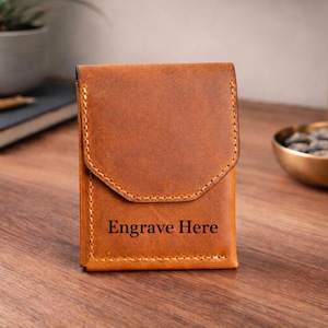 May include: A brown leather wallet with a flap closure. The wallet has a stitched border and the words "Engrave Here" are printed on the front. The wallet is sitting on a wooden surface.