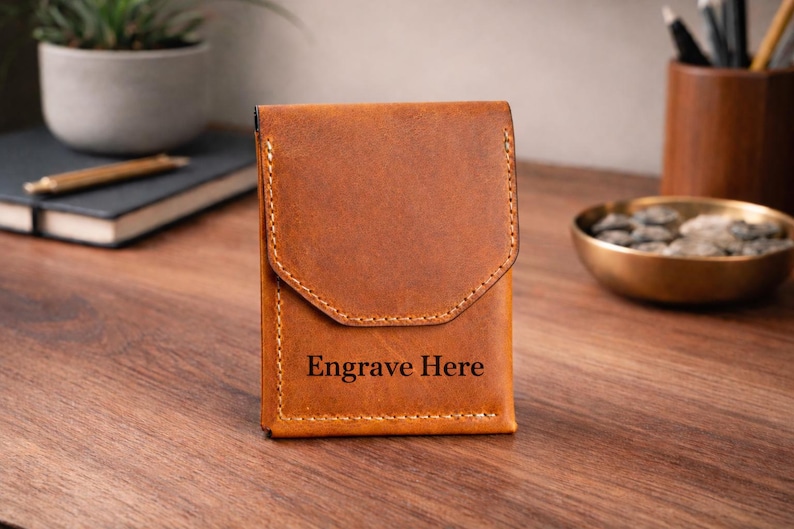 May include: A brown leather wallet with a flap closure. The wallet has a stitched border and the words "Engrave Here" are printed on the front. The wallet is sitting on a wooden surface.