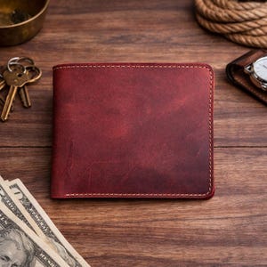 May include: A burgundy leather wallet with white stitching. The wallet is rectangular and appears to be bi-fold. The background includes keys, a watch, and US currency on a wooden surface. The wallet is the focal point of the image.