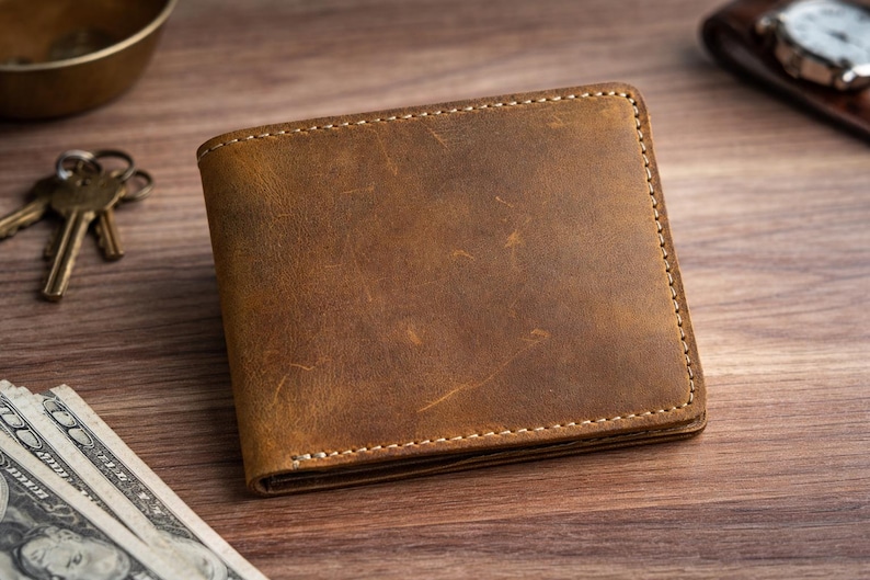 May include: A brown leather wallet with visible stitching and a worn texture. The wallet is rectangular and appears to be new. Keys, a small bowl, and cash are visible in the background.