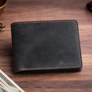 May include: A dark gray leather wallet with visible stitching. The wallet is rectangular and appears to be new. Other items on the wooden surface include keys, cash, and a watch.