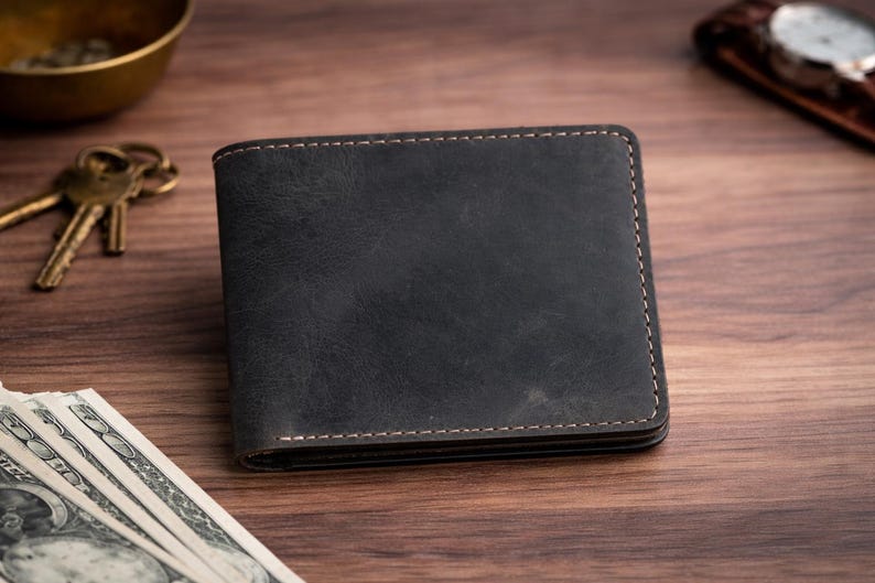 May include: A dark gray leather wallet with visible stitching. The wallet is rectangular and appears to be new. Other items on the wooden surface include keys, cash, and a watch.